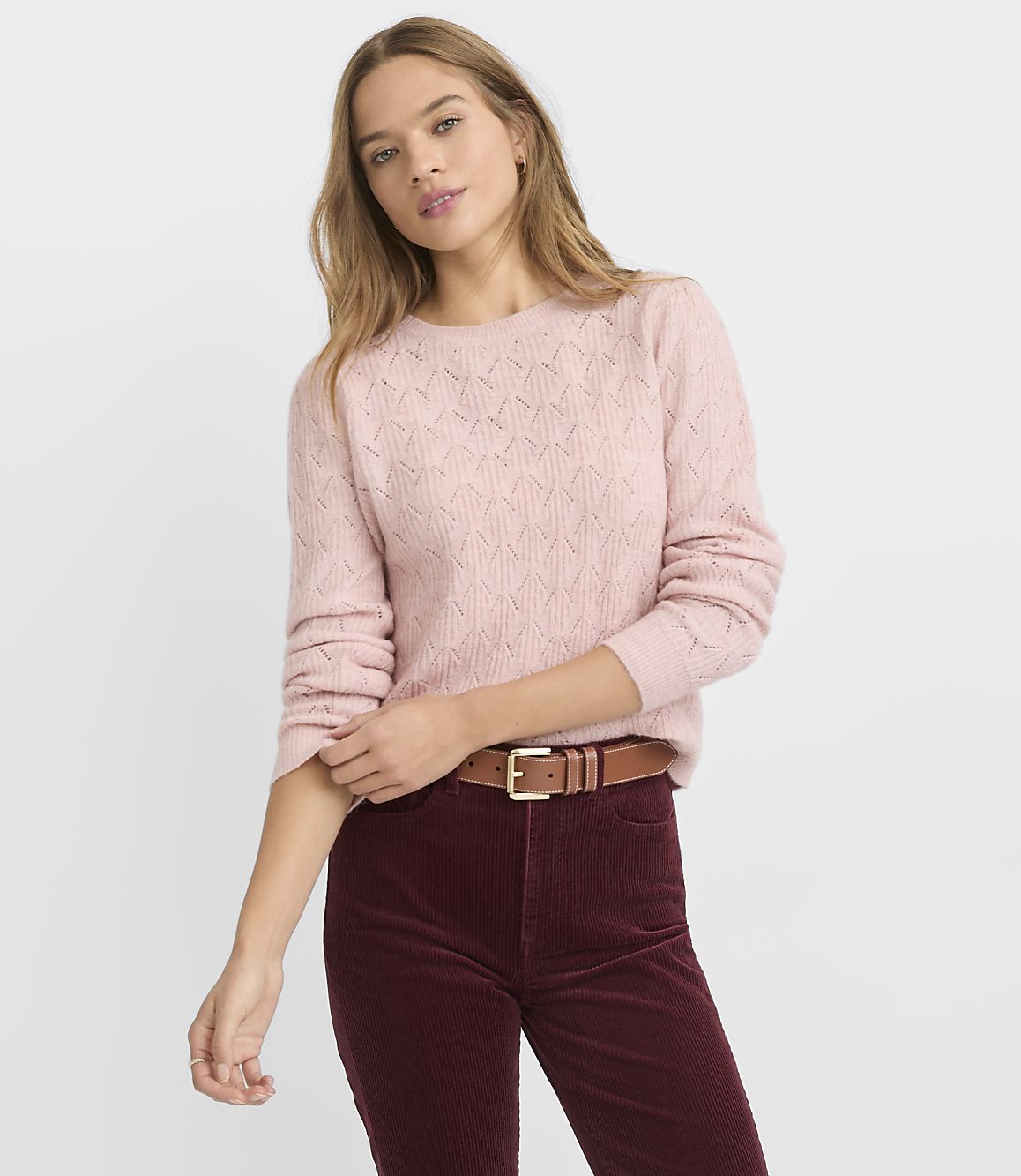 A young woman in a pink sweater and burgundy pants stands against a plain gray background.