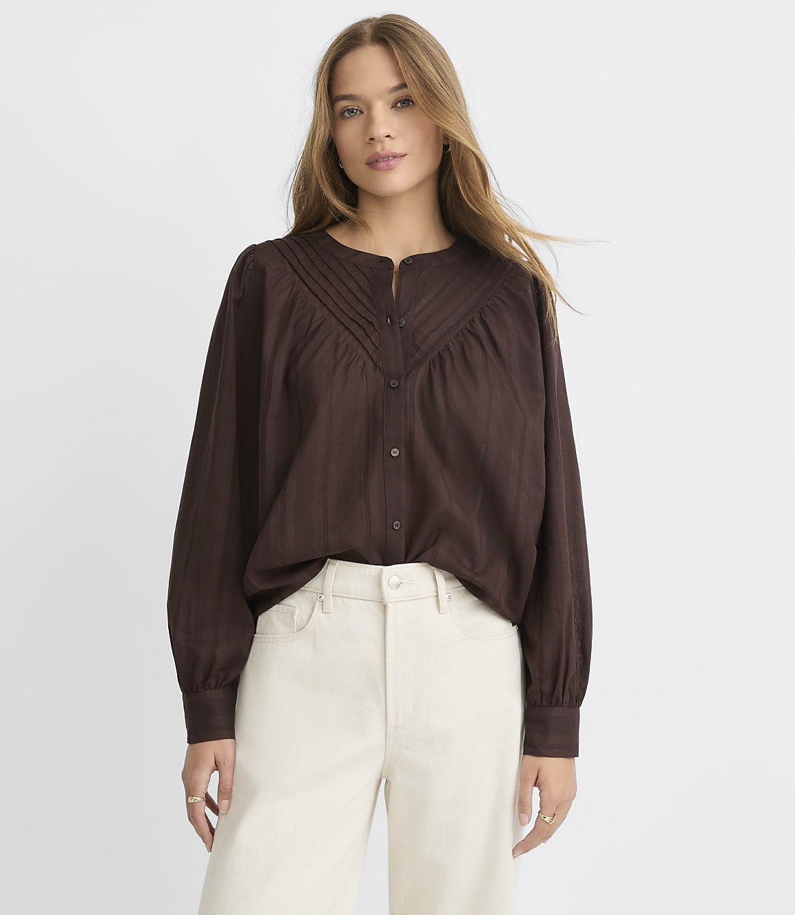 A woman in a brown blouse and cream pants stands against a plain white background.
