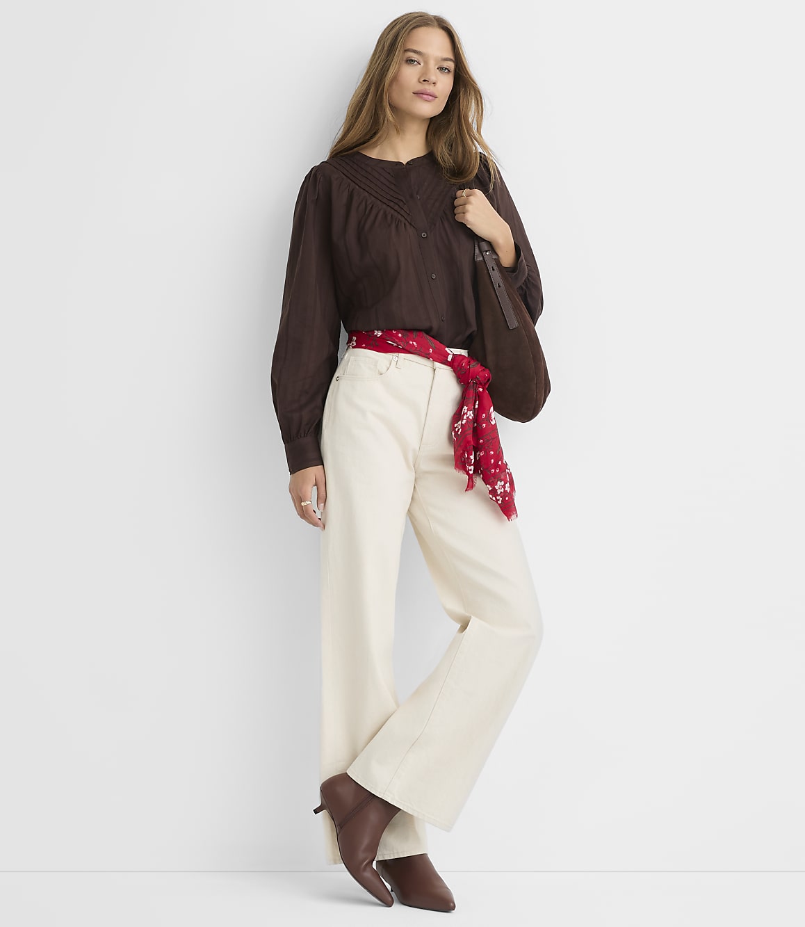A woman in a brown blouse, cream pants, and a red scarf stands against a plain white background.