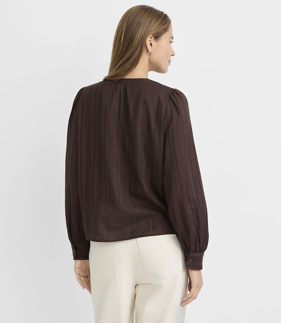 A woman in a brown blouse and cream pants stands against a plain white background.