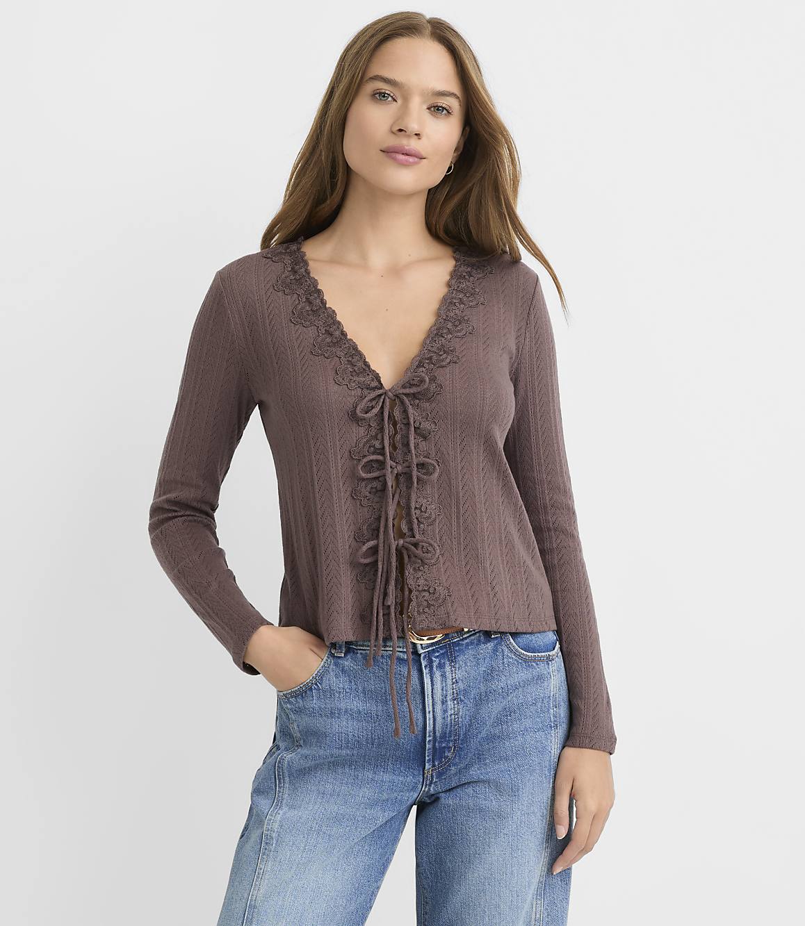 A woman in a brown lace-up cardigan and blue jeans stands against a plain gray background.