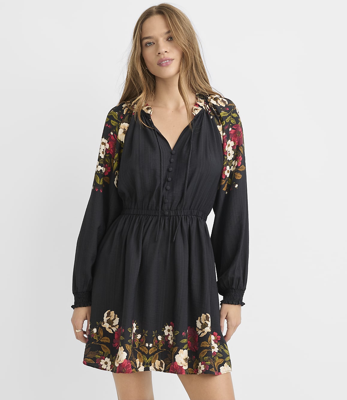 A woman in a black floral-accented dress stands against a plain white background.