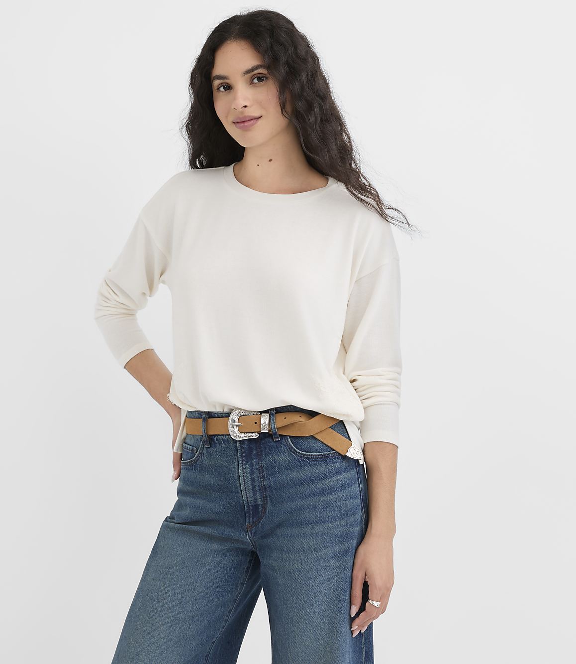 A woman in a cream sweater and blue jeans stands against a plain white background.