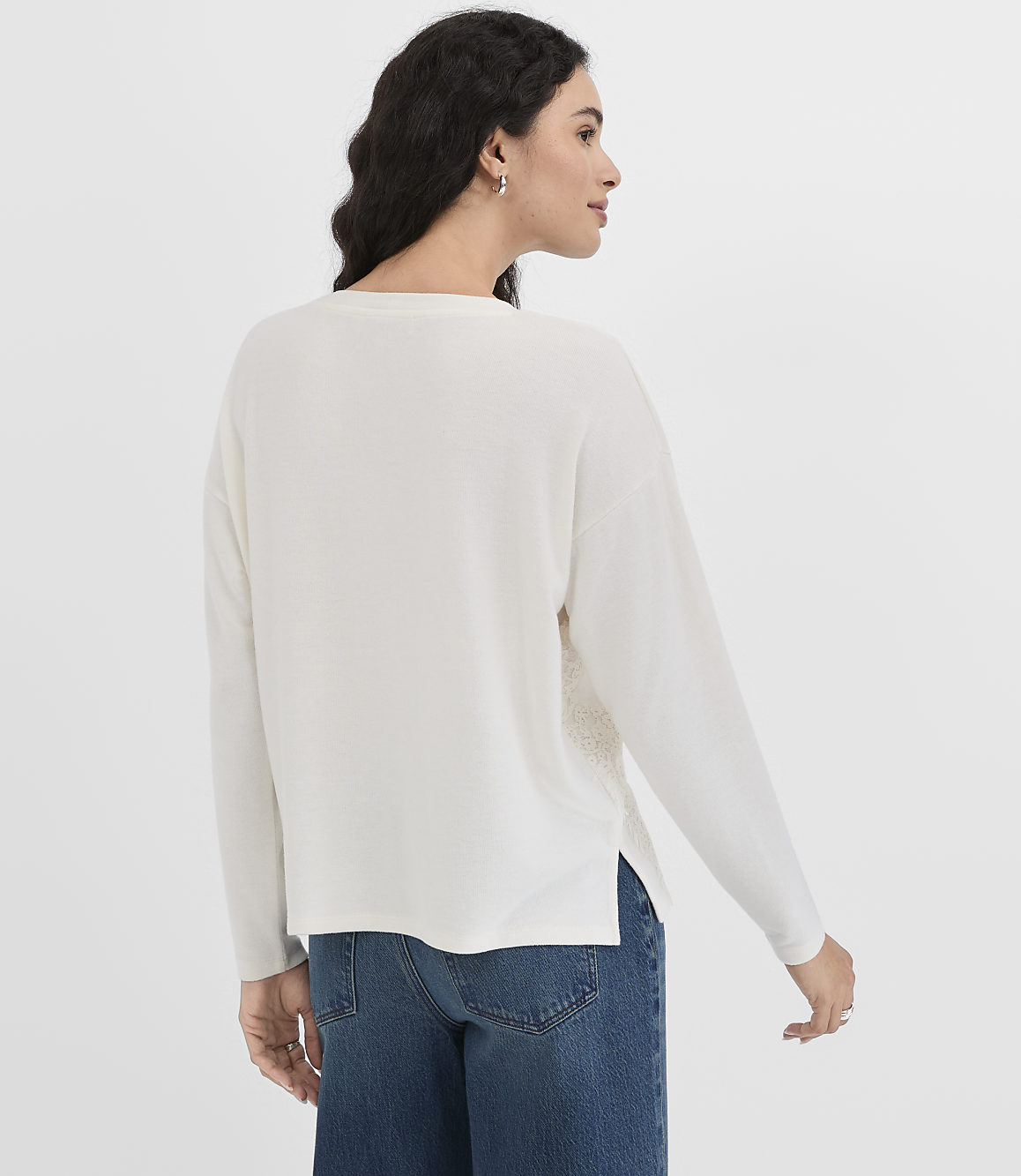 A woman in a white sweater and blue jeans stands against a plain gray background.