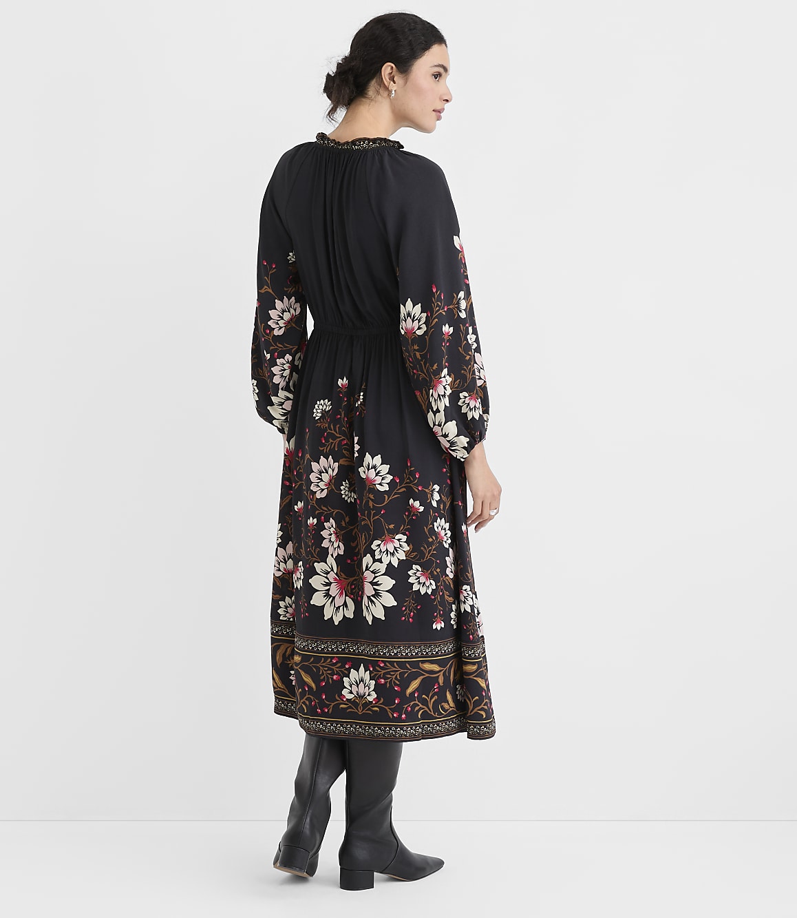 A woman in a black floral dress and black boots stands against a plain white background.