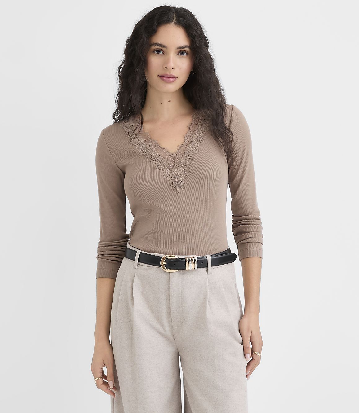 A woman in a brown lace-trimmed top and light-colored pants stands against a plain white background.