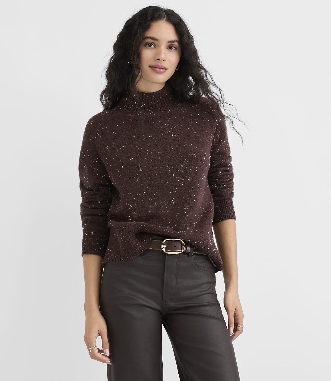A woman in a speckled brown turtleneck sweater and dark pants stands against a plain gray background.