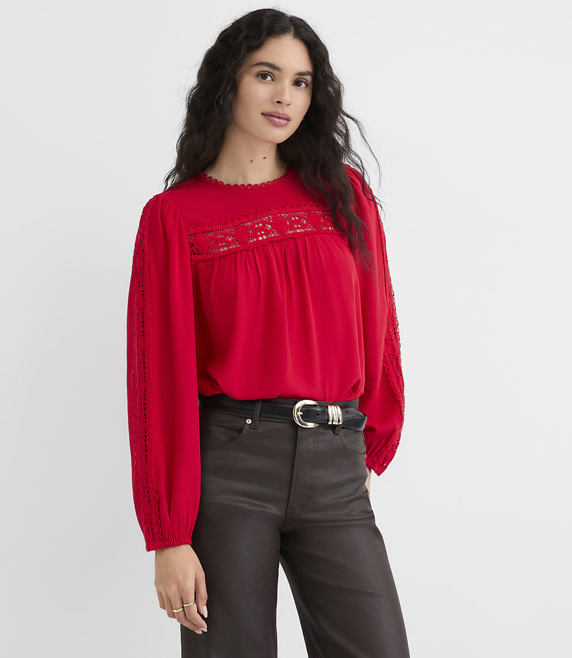 A woman in a red lace-trimmed blouse and dark pants stands against a plain red background.