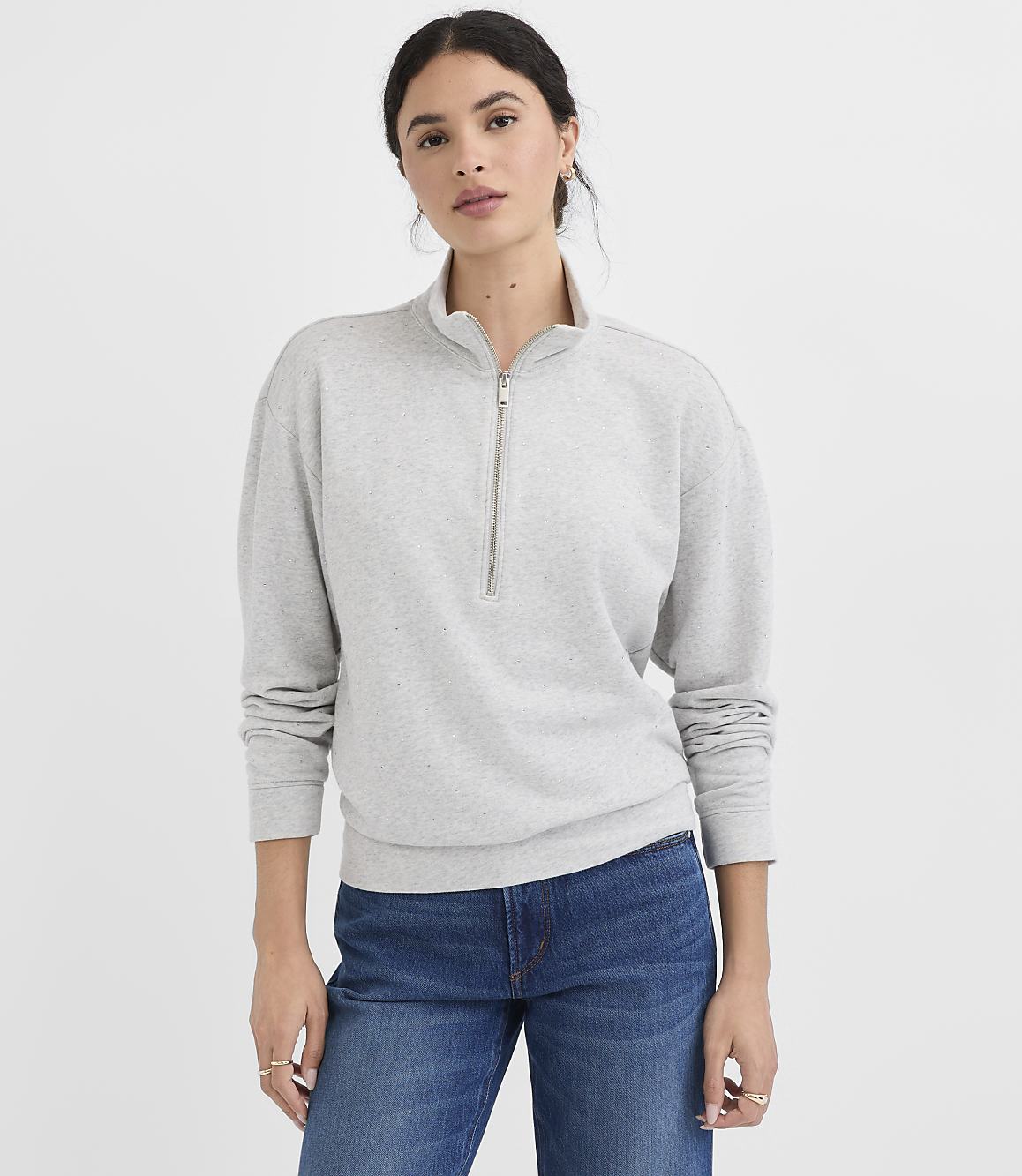 A woman in a gray quarter-zip sweatshirt and blue jeans stands against a plain white background.