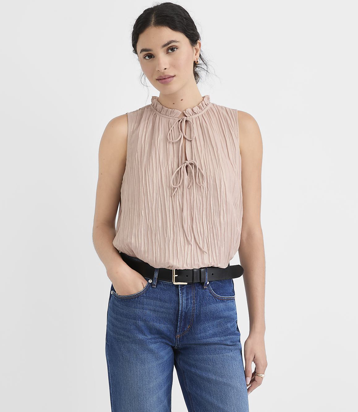 A woman in a ruffled pink top and blue jeans stands against a plain, light background.