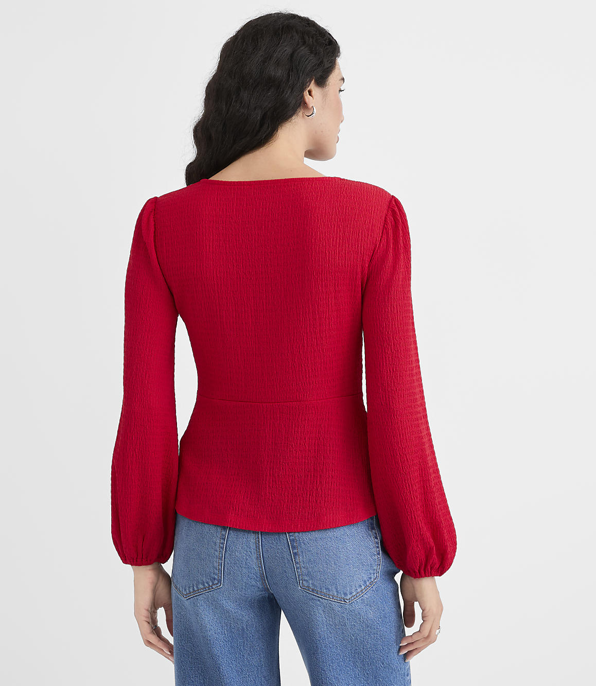 A woman in a red textured top and blue jeans stands against a plain white background.