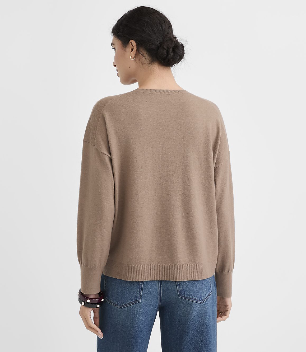 A woman in a tan sweater and blue jeans stands against a plain white background.