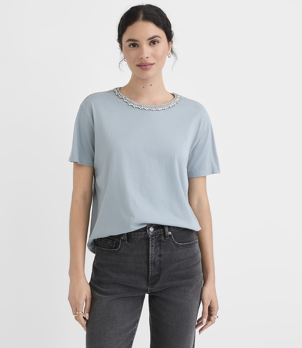A woman in a light blue t-shirt with a decorative neckline and dark jeans stands against a plain white background.