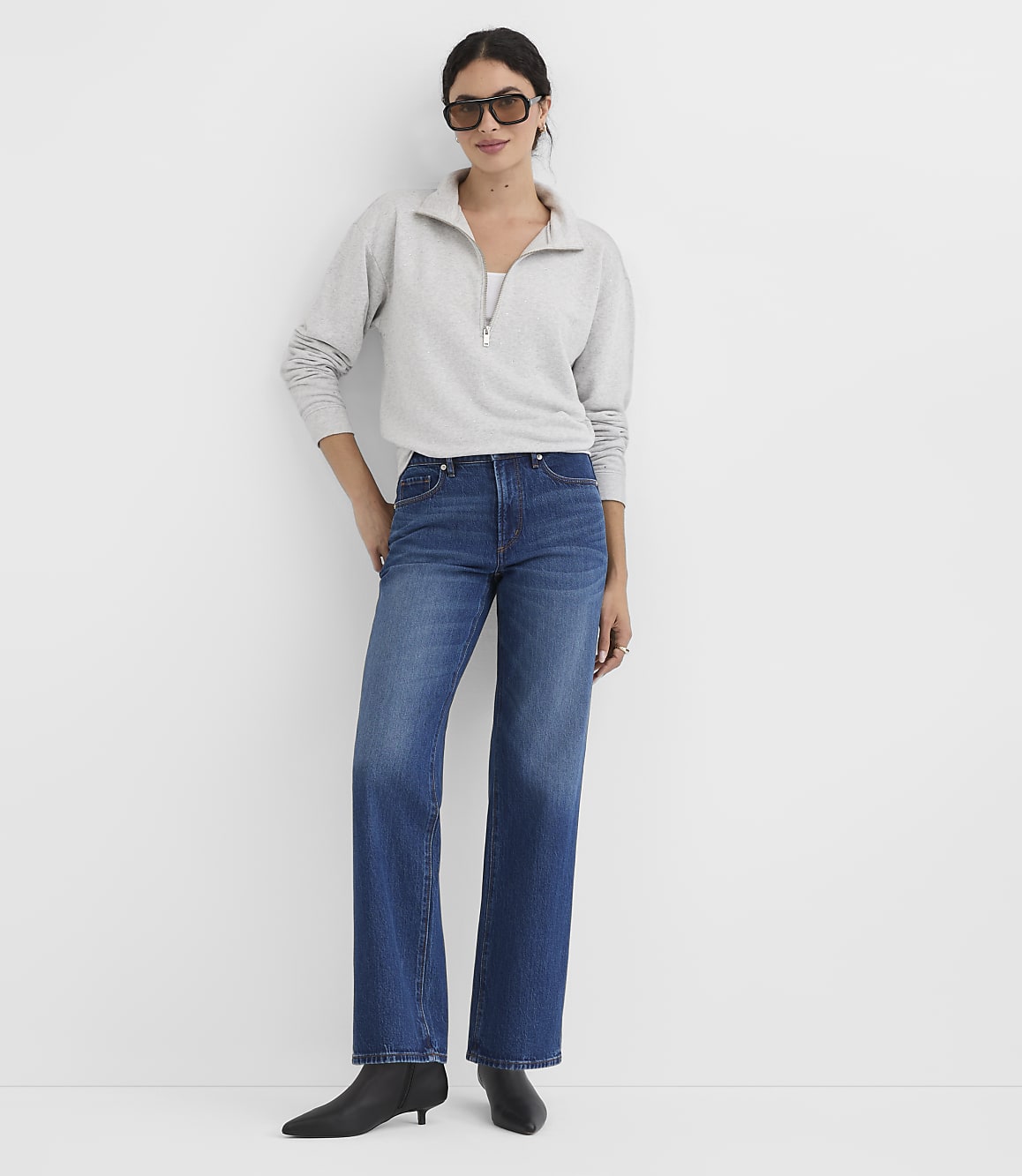 A woman in a grey quarter-zip sweatshirt and blue jeans stands against a plain white background.