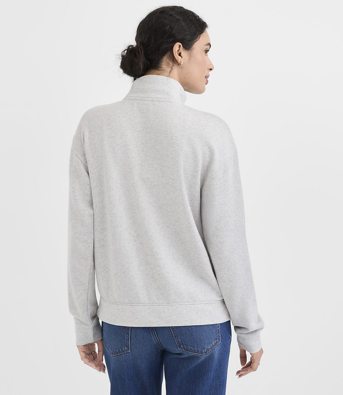 A woman in a grey quarter-zip sweatshirt and blue jeans stands against a plain white background.