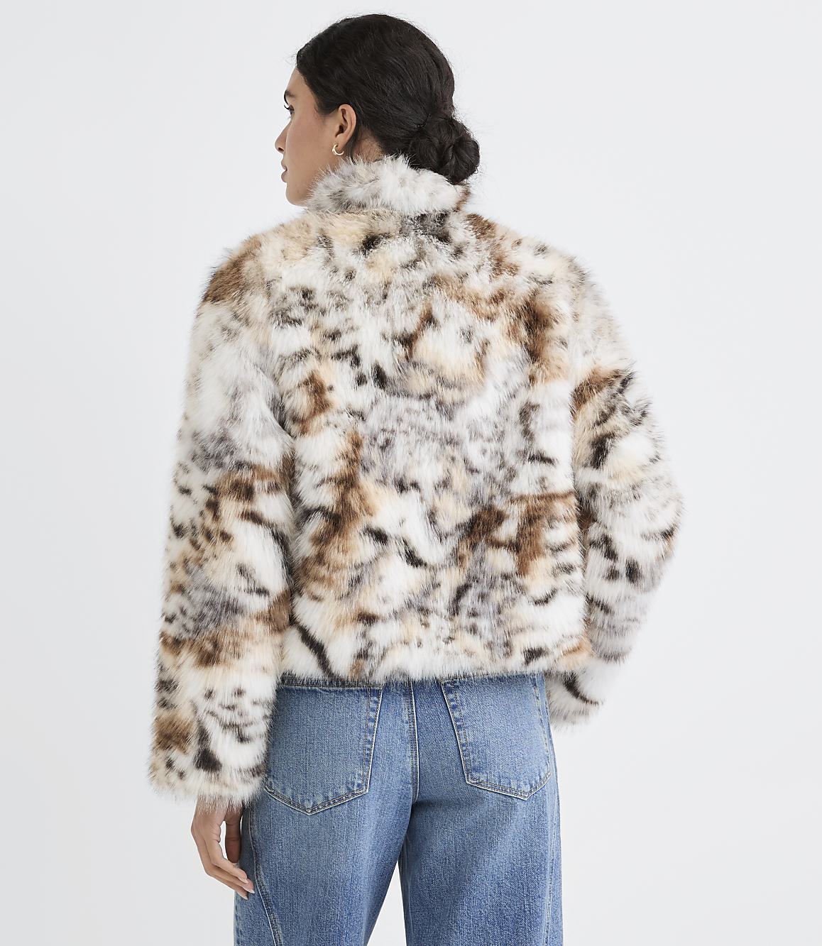 A woman in a spotted faux fur jacket and blue jeans stands against a plain white background.
