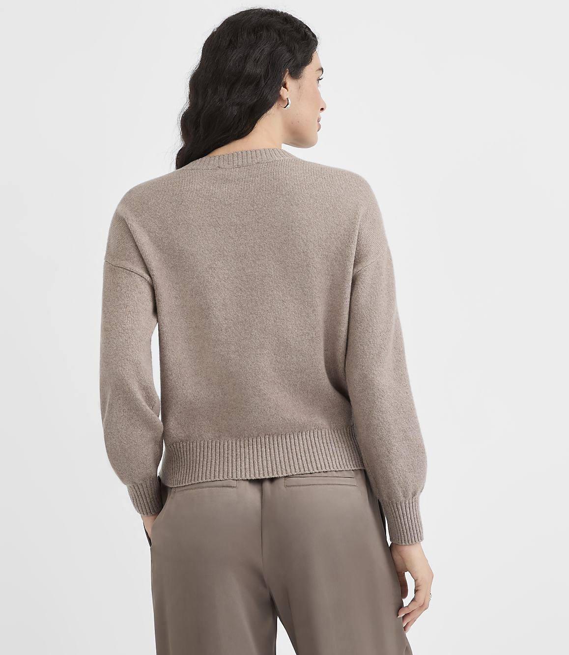 A woman in a taupe sweater and pants stands against a plain white background.