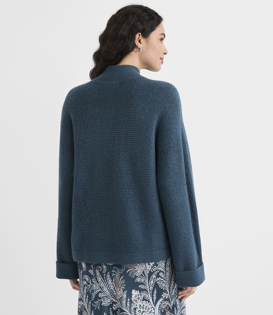 A woman in a teal sweater and patterned skirt stands against a plain white background.