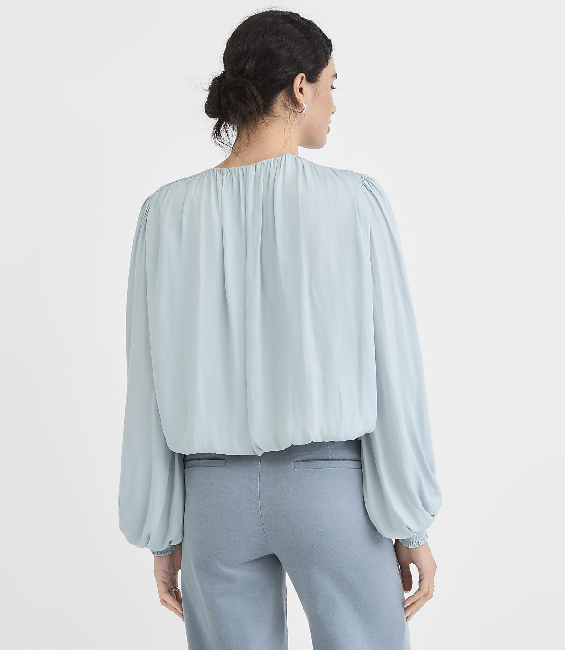 A woman in a light blue blouse and blue pants stands against a plain white background.