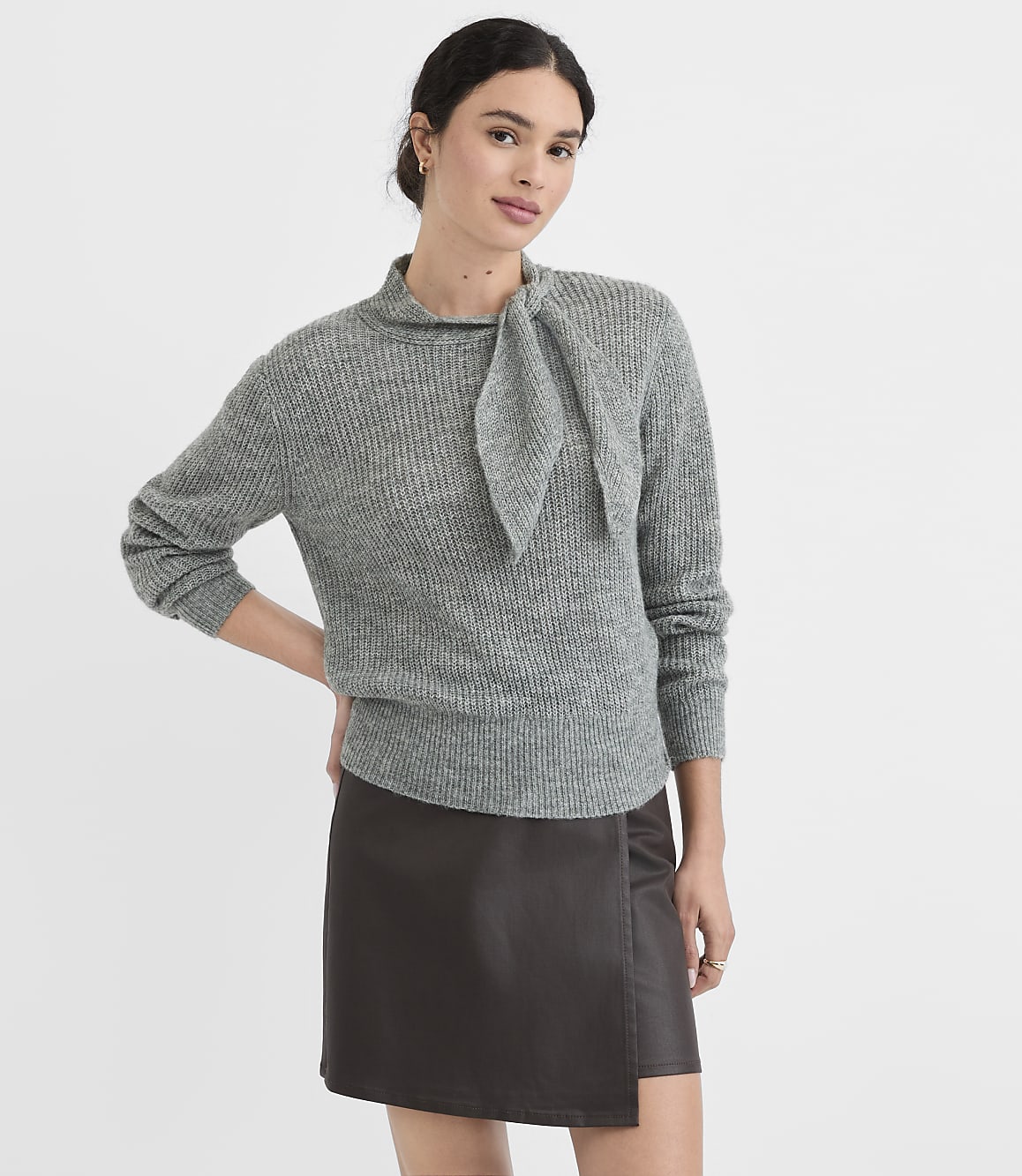 A woman in a grey sweater and dark skirt stands against a plain white background.
