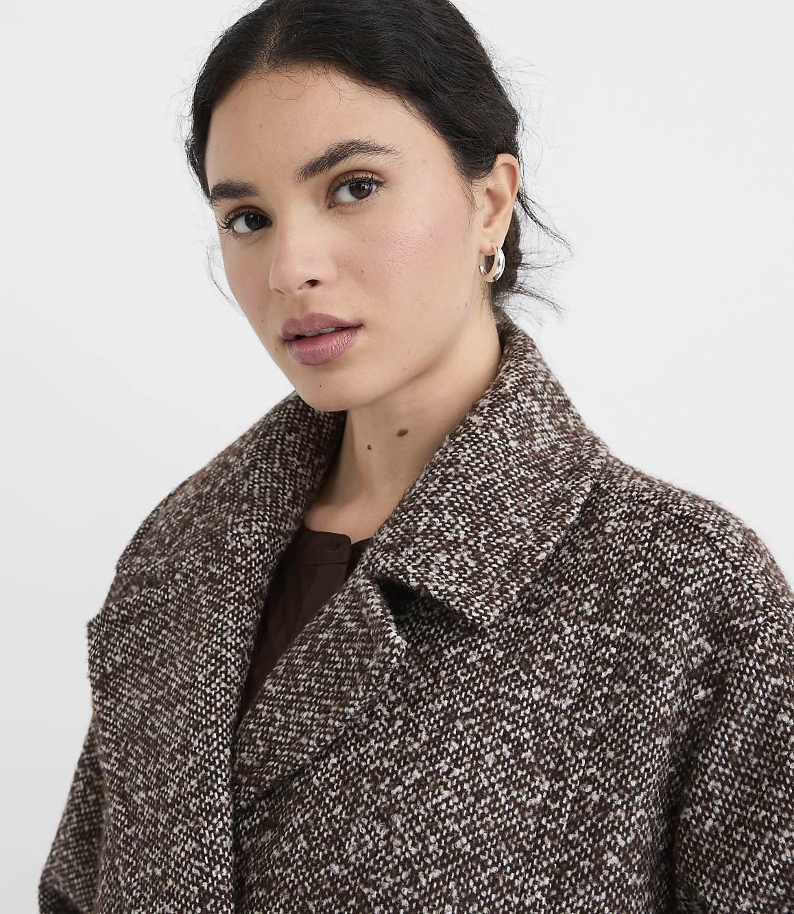 A woman in a textured brown coat with a white flecked pattern looks forward against a plain white background.
