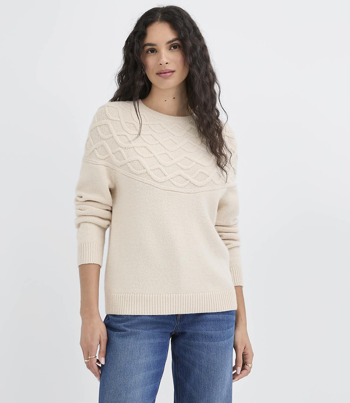 A woman in a cream-colored sweater and blue jeans stands against a plain white background.