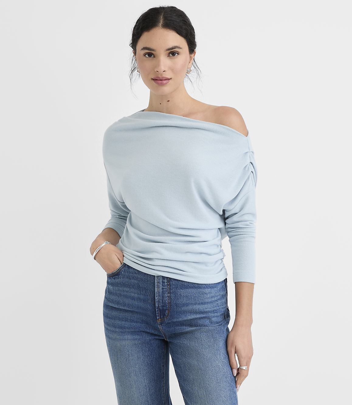 A woman in a light blue off-the-shoulder top and jeans stands against a plain white background.