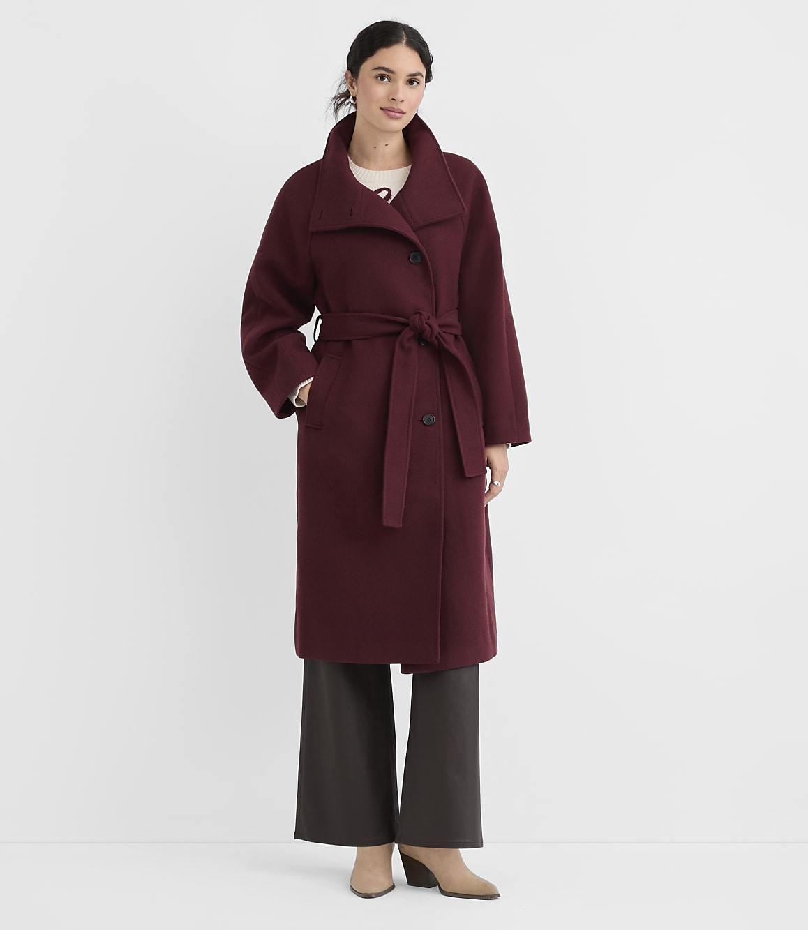 A woman in a burgundy coat and dark pants stands against a plain white background.