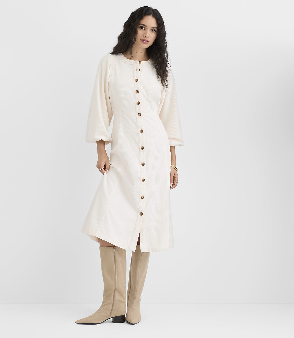 A woman in a cream-colored button-down dress and beige boots stands against a plain white background.