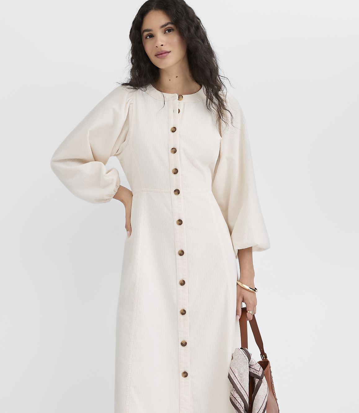 A woman in a cream-colored button-down dress with voluminous sleeves stands against a plain white background, holding a brown leather tote bag.