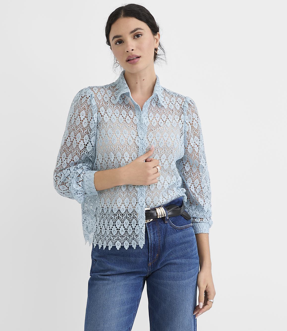 A woman in a light blue lace blouse and jeans stands against a plain white background.