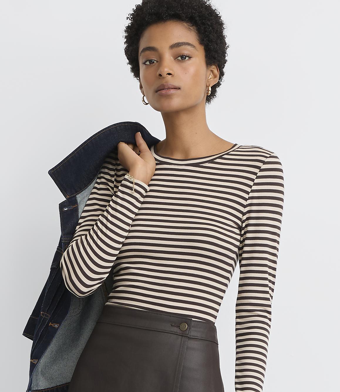 A woman in a striped shirt and leather skirt poses with a denim jacket draped over her shoulder against a plain background.