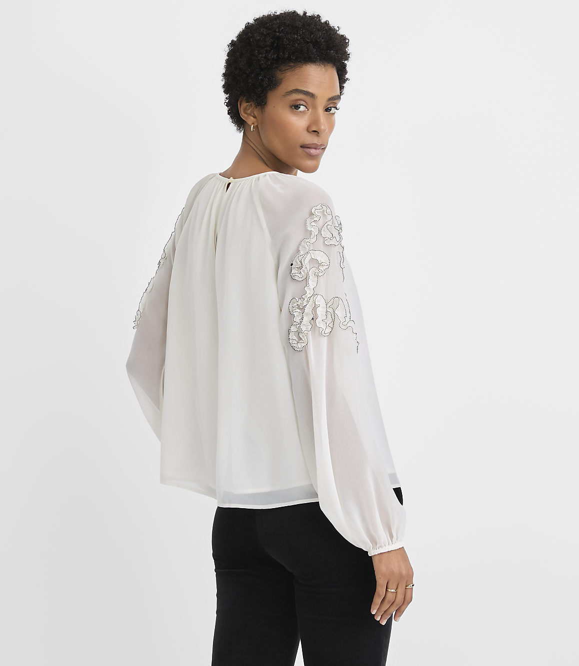 A woman in a white embellished blouse and black pants stands against a plain white background.