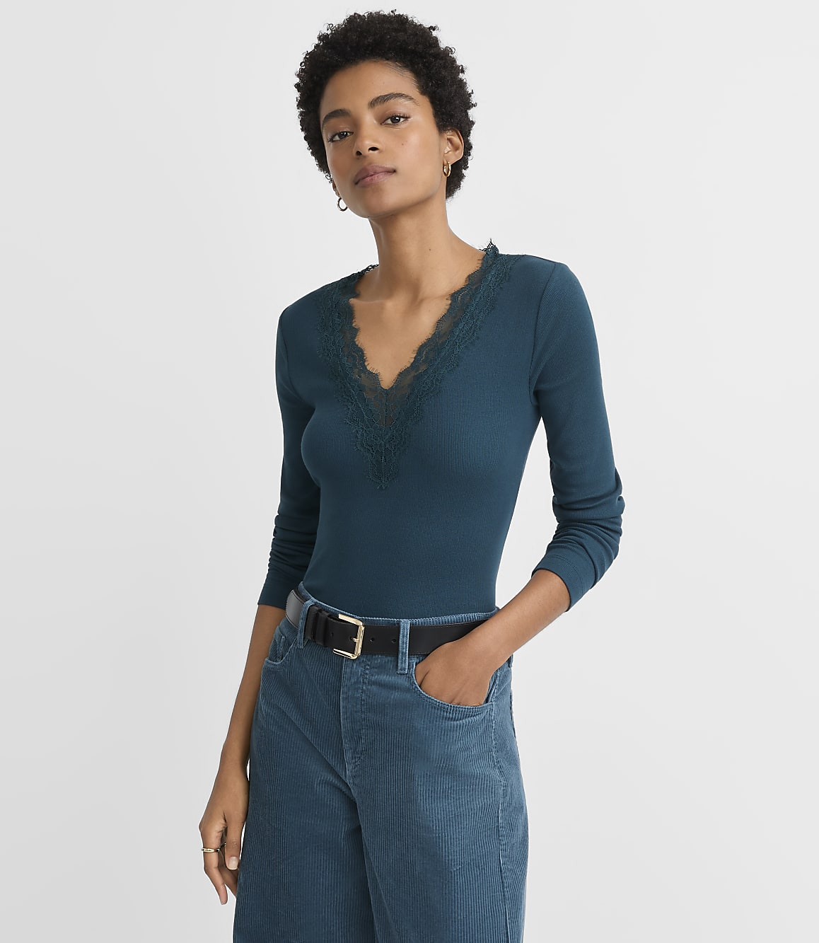 A woman in a teal ribbed long-sleeve shirt with lace trim and teal corduroy pants stands against a plain gray background.
