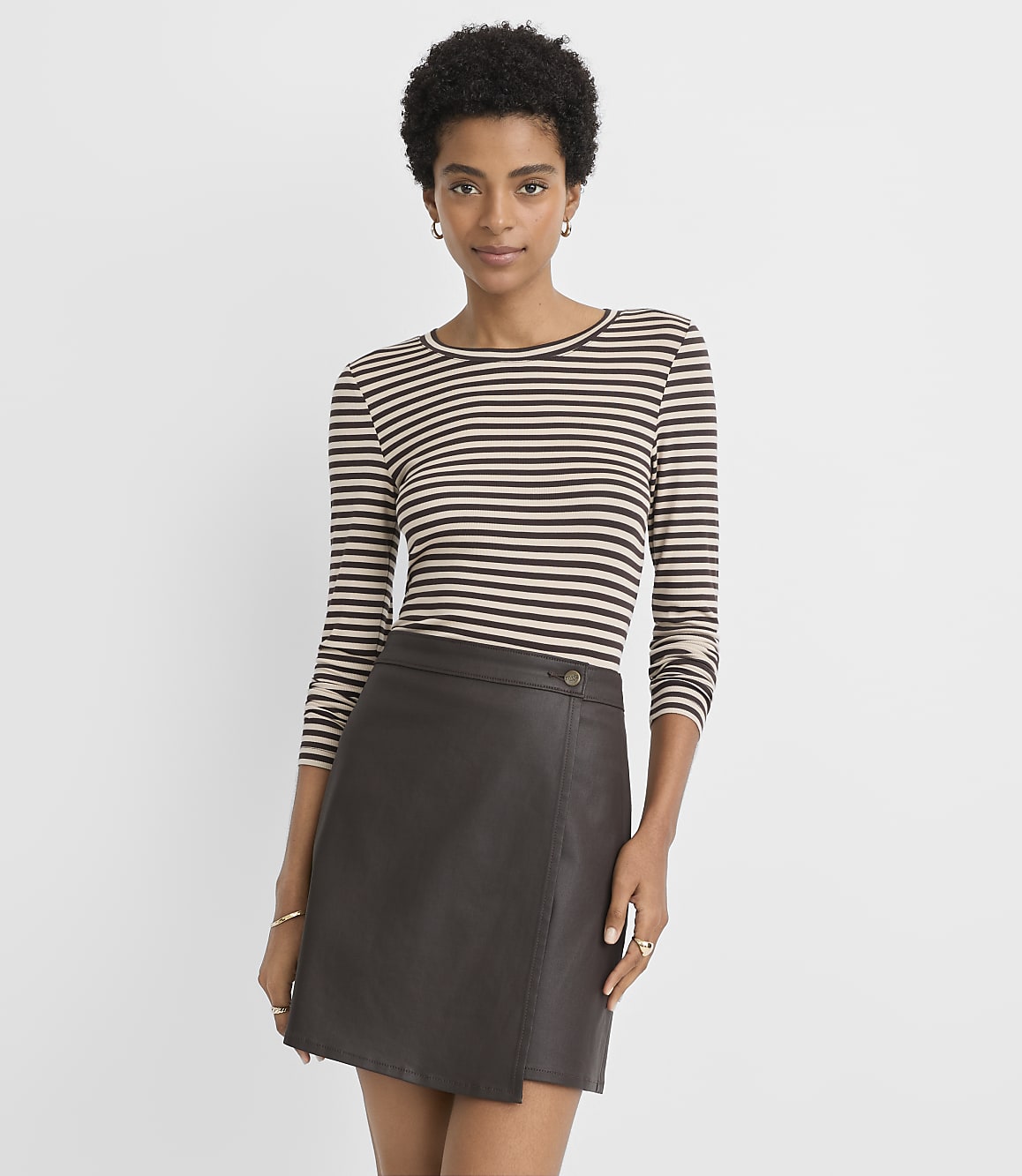A woman in a striped shirt and a brown leather skirt stands against a plain, light gray background.