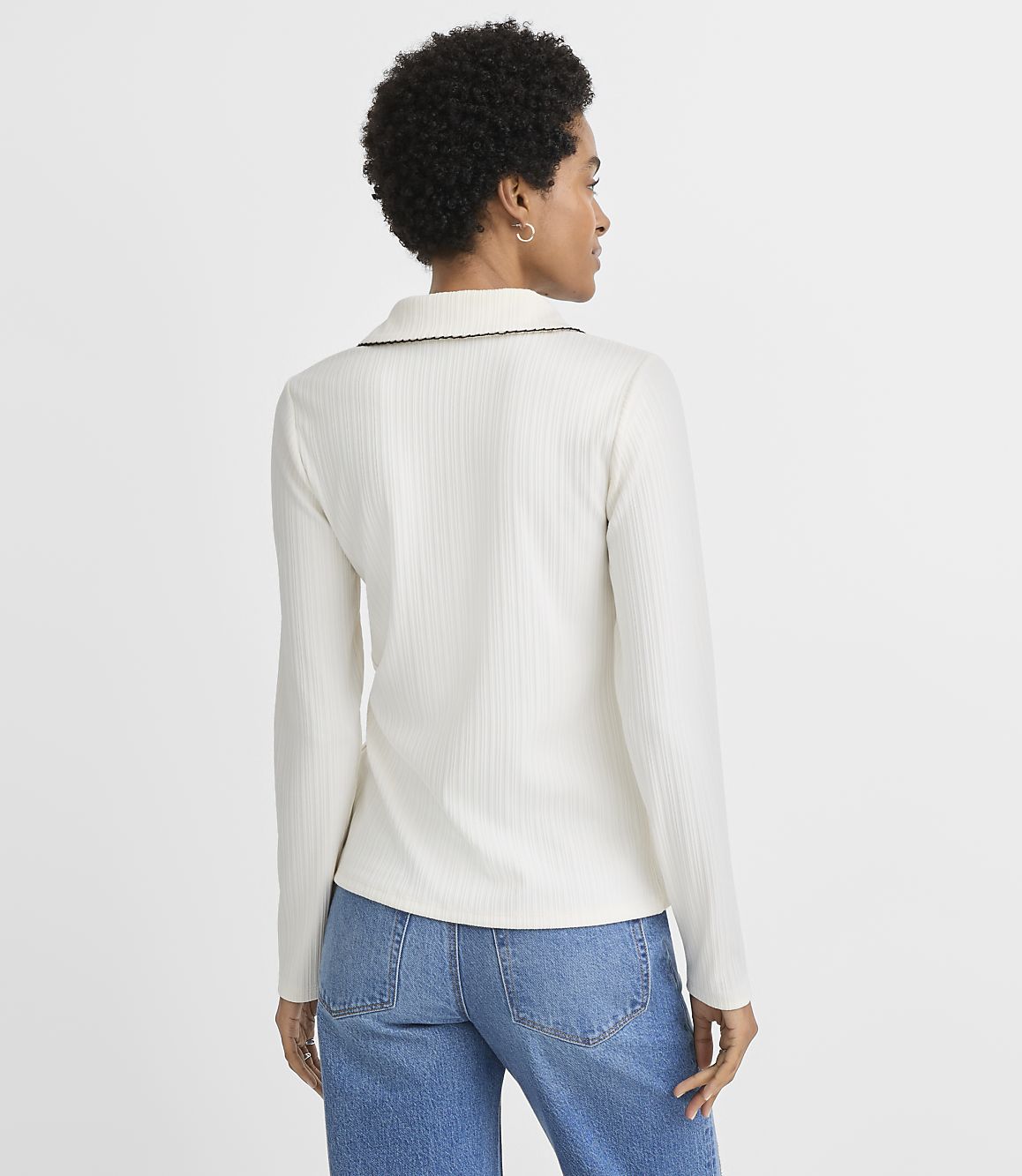 A woman in a white ribbed long-sleeved shirt and blue jeans stands against a plain white background.