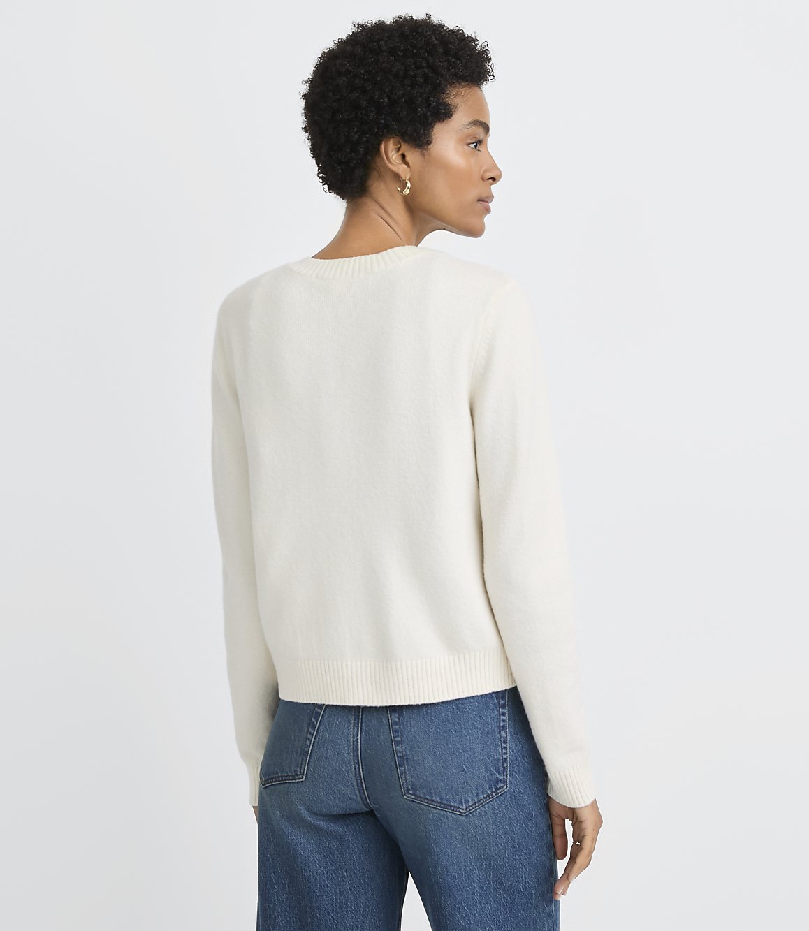 A woman in a cream sweater and blue jeans stands against a plain white background.