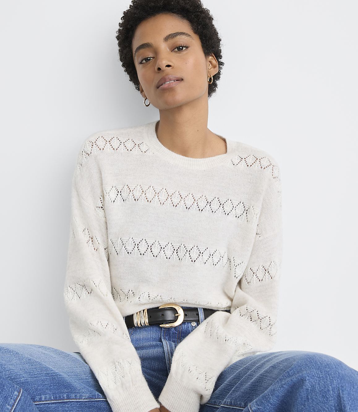 A woman in a cream-colored, open-knit sweater and blue jeans sits against a plain white background.