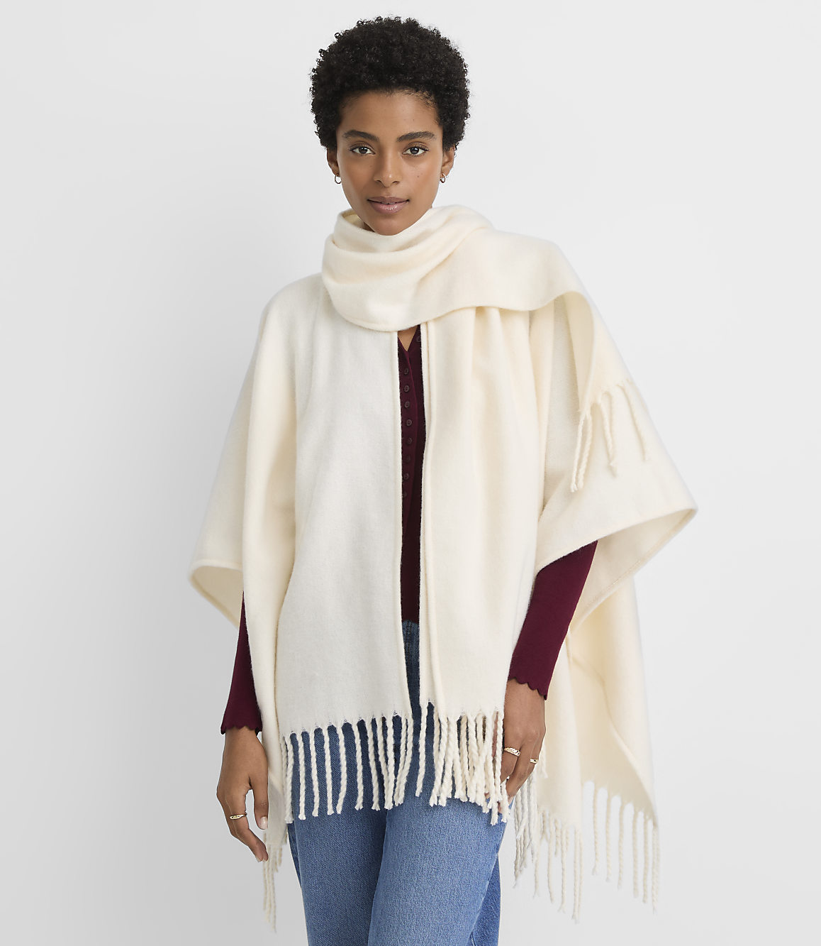 A woman in a cream-colored poncho with fringe stands against a plain white background.