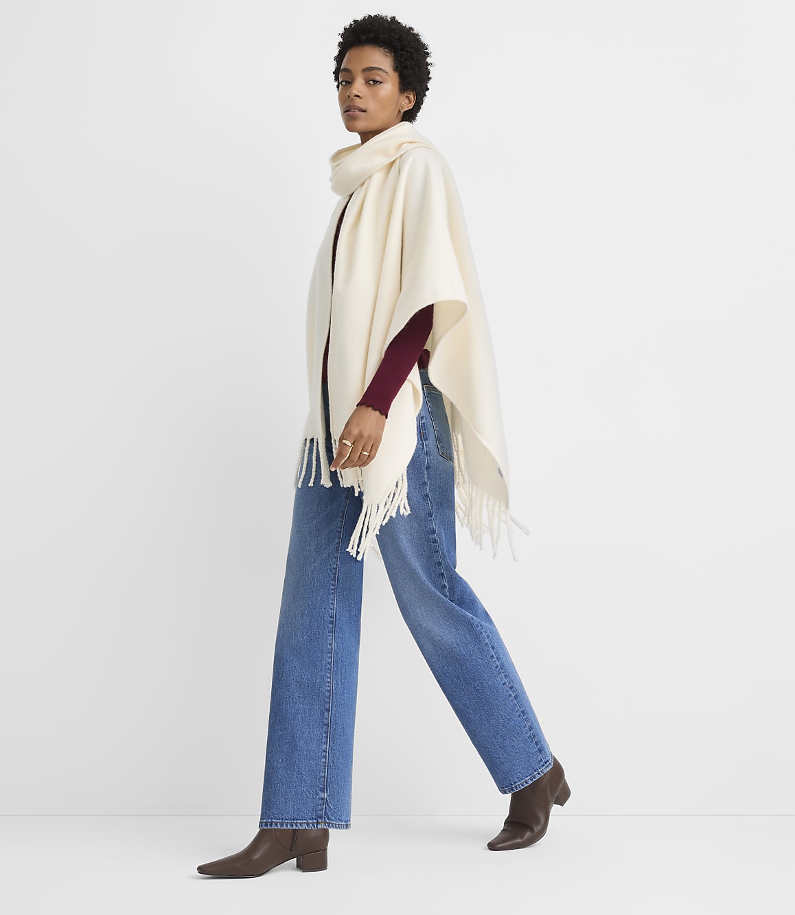 A woman in a cream-colored poncho and jeans walks against a plain white background.