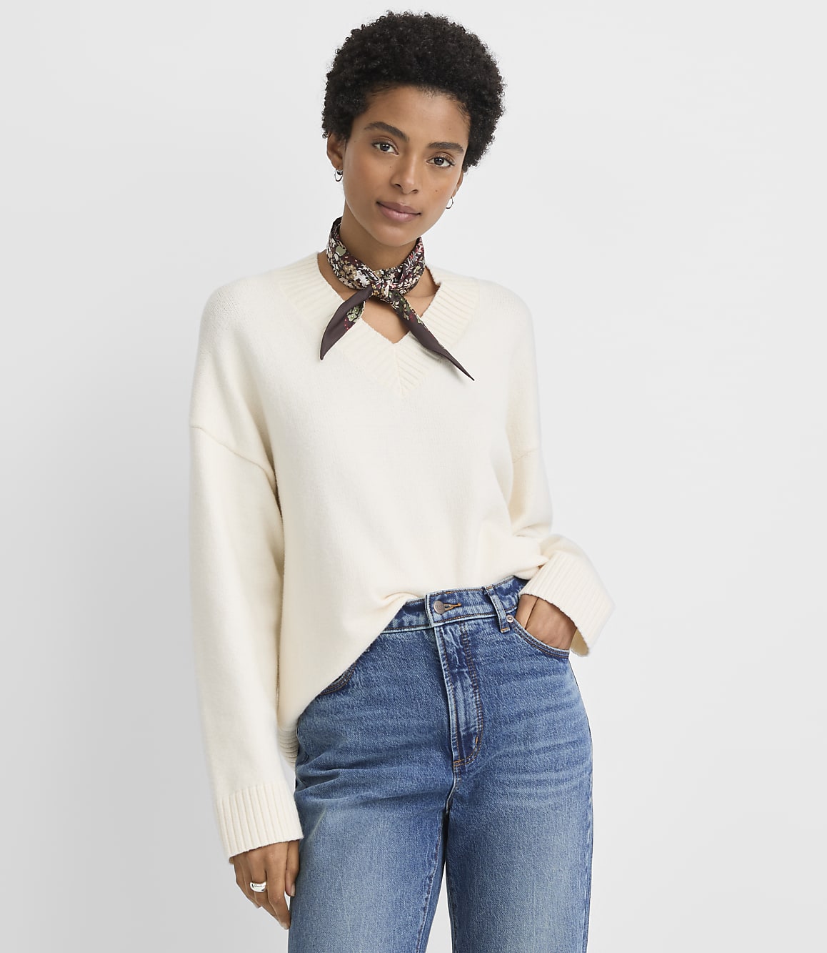 A woman in a cream v-neck sweater and blue jeans stands against a plain white background.