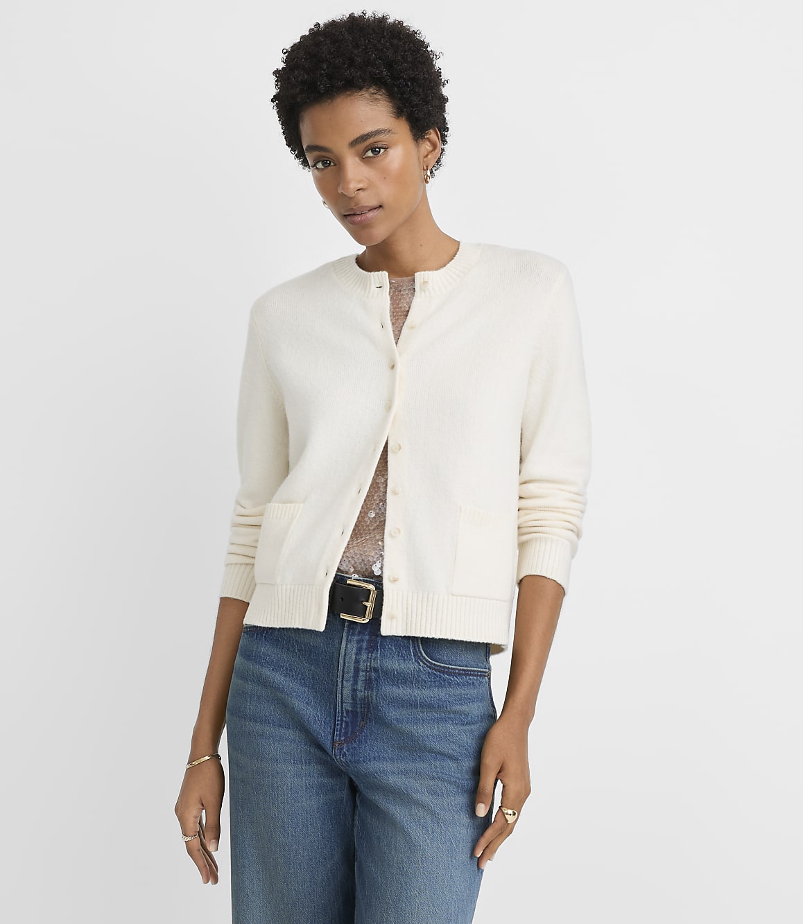 A woman in a cream cardigan and blue jeans stands against a plain white background.