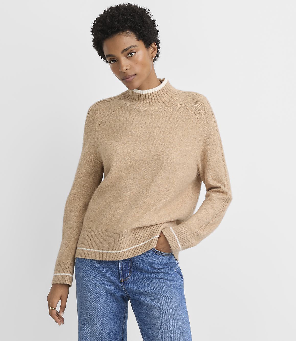 A woman in a tan mock-neck sweater and blue jeans stands against a plain gray background.