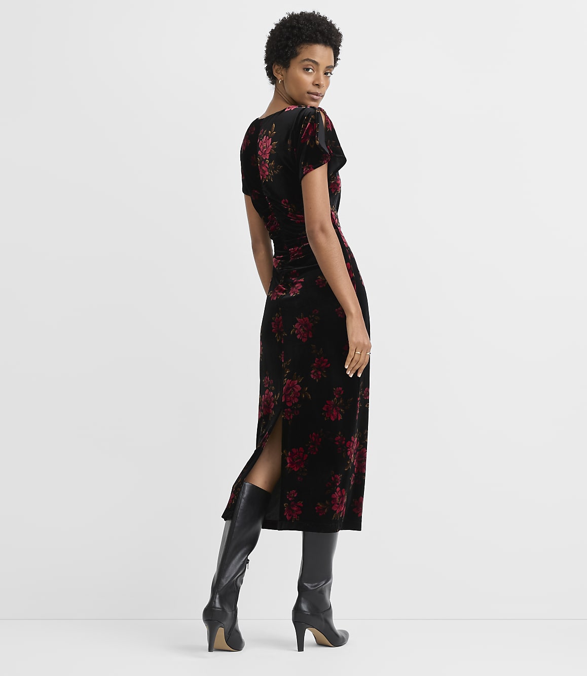 A woman in a black velvet dress with red floral print stands against a plain gray background.