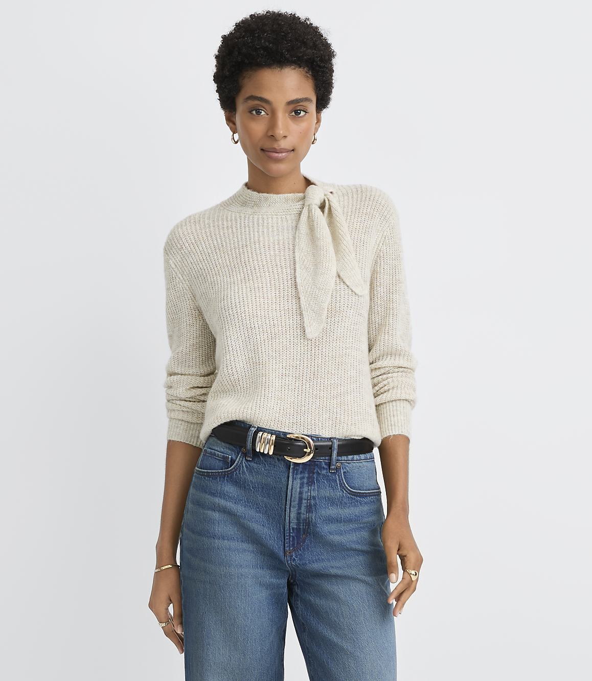 A woman in a cream-colored sweater with a bow detail and blue jeans stands against a plain, light gray background.