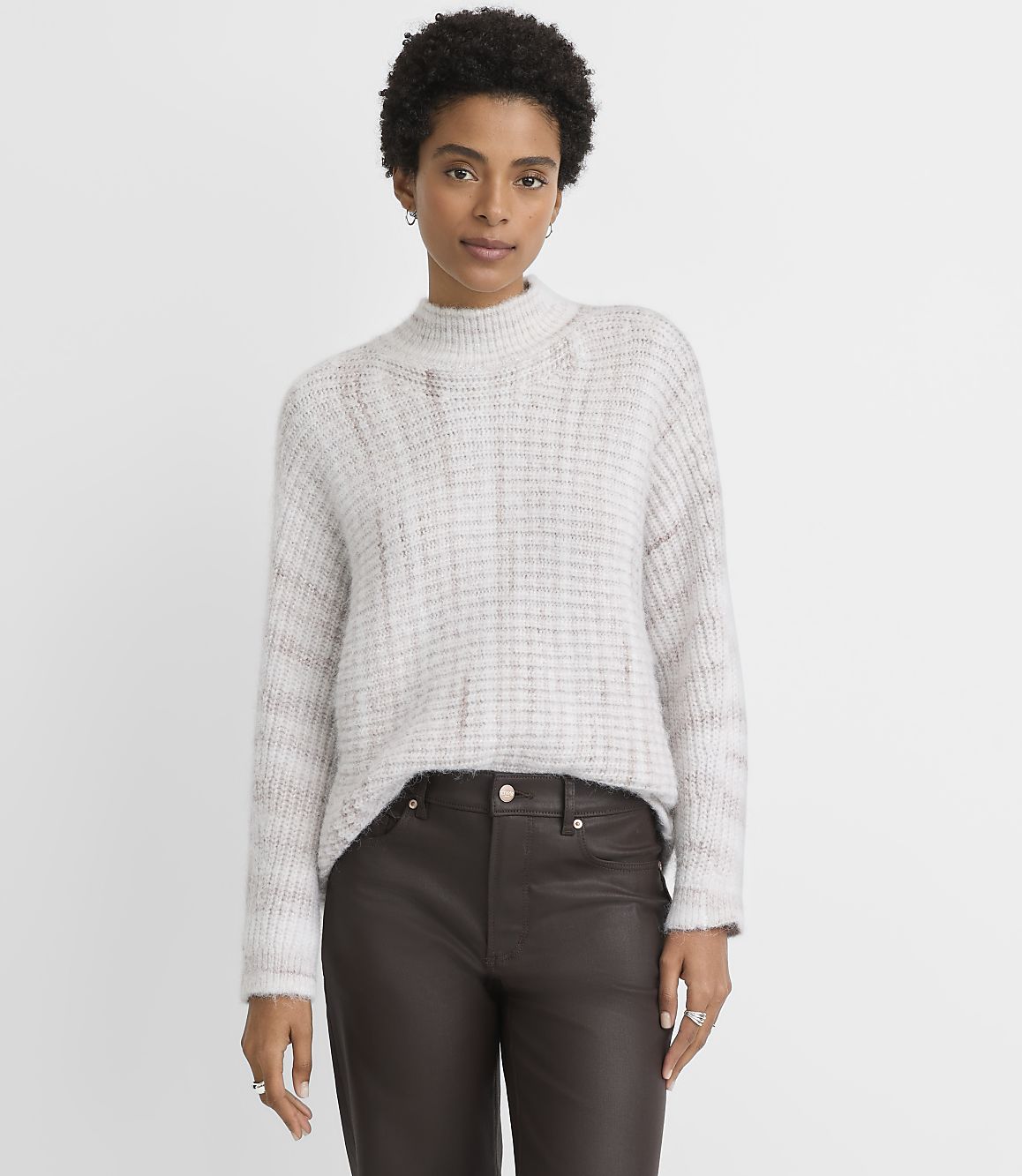 A woman in a light-colored, textured sweater and dark pants stands against a plain, light gray background.