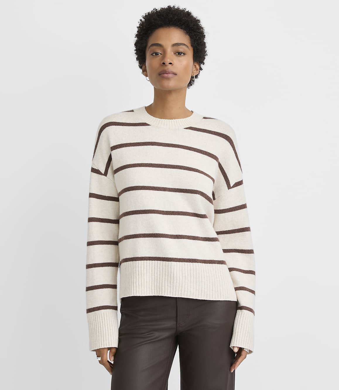 A woman in a cream and brown striped sweater and dark pants stands against a plain white background.
