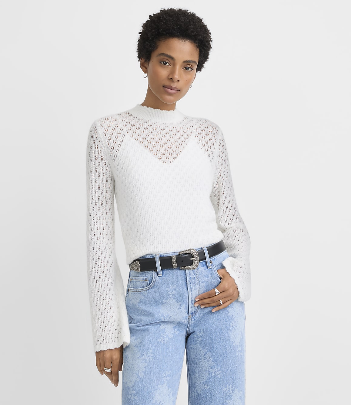 A woman in a white crochet sweater and patterned jeans stands against a plain white background.