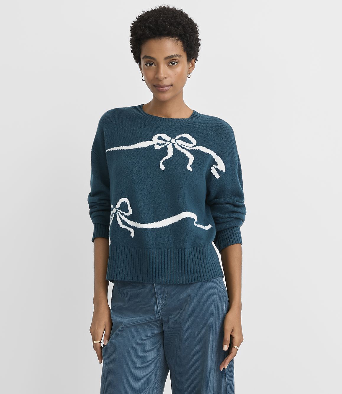 A woman in a teal sweater with white ribbon designs stands against a plain gray background.