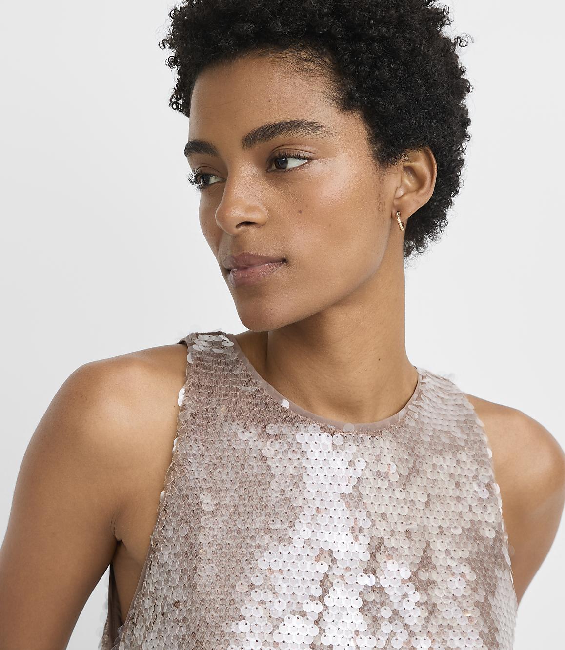 A woman with short, dark hair and a sequined top is shown against a plain, light gray background.